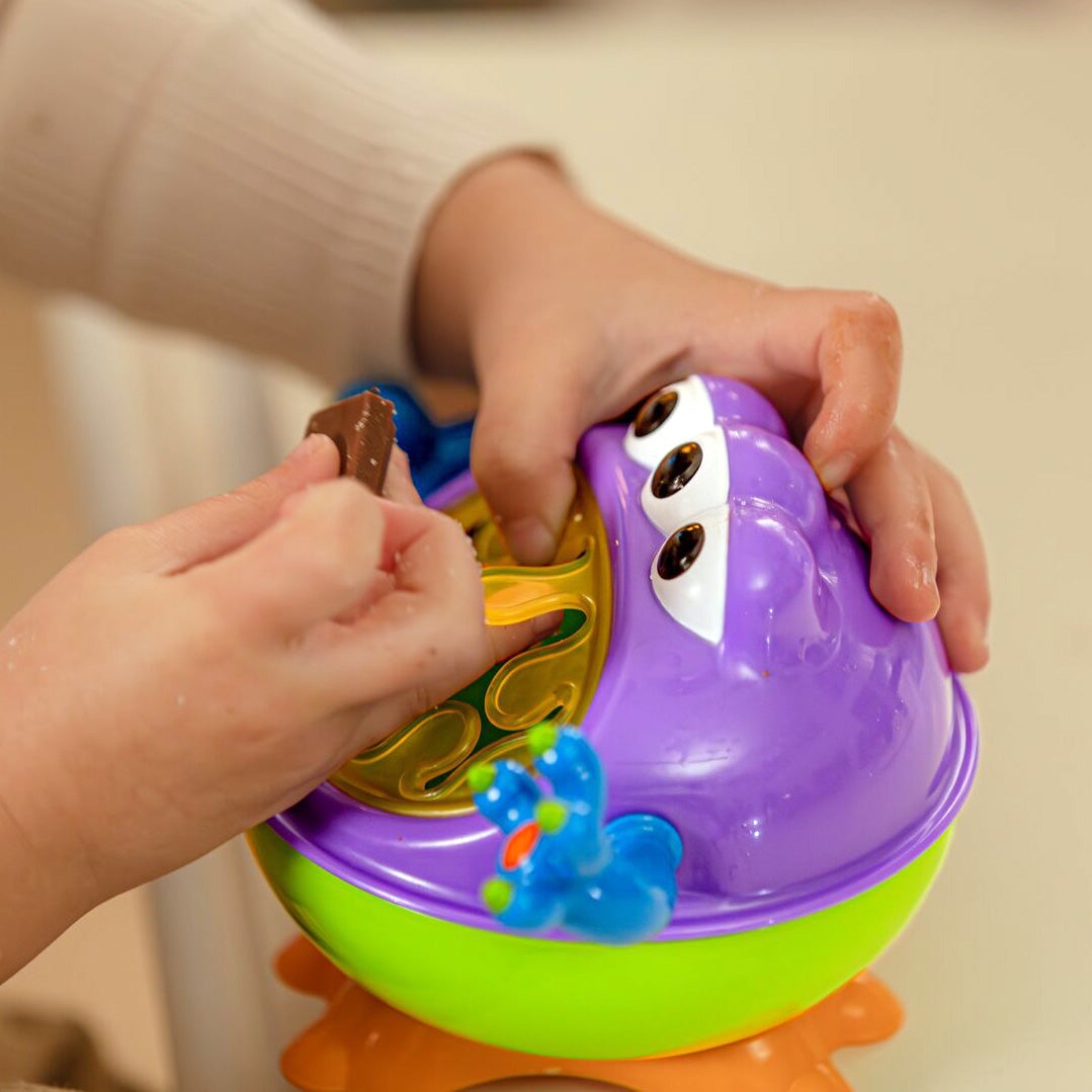Gros plan de mains d’enfant prenant une collation dans le porte-collation Nûby 3D Monster violet, vert, bleu et orange.